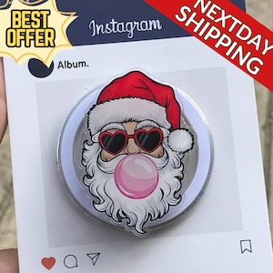 May include: A round, silver pop socket featuring a cartoon Santa Claus with heart-shaped sunglasses blowing a pink bubble. Santa wears a red hat and has a white beard. The background includes Instagram-themed elements.