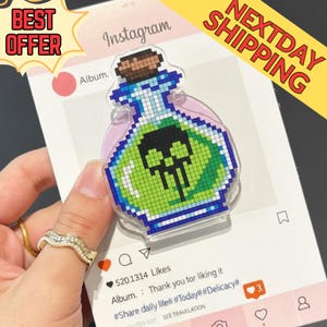 May include: A clear acrylic potion bottle-shaped charm with a pixelated design. The bottle contains green liquid and a black skull. The charm is on a social media-themed background with the text "Instagram" and "Album."