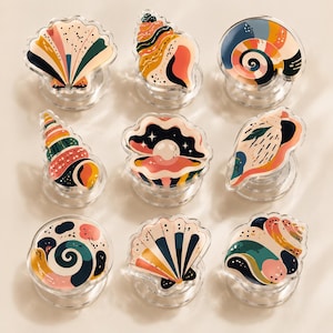 May include: A collection of nine circular phone grips featuring various seashell designs in a palette of cream, pink, teal, and dark blue. Each grip displays a unique shell shape, including fan, spiral, and conch styles, with clear plastic bases.
