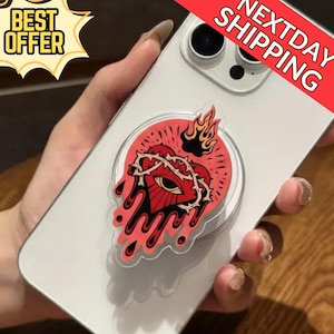 Puede incluir: Un soporte transparente para teléfono con un diseño de corazón rojo que presenta una corona de espinas, un ojo y llamas. El soporte está adherido a un teléfono blanco. Se ven las palabras "BEST OFFER" y "NEXTDAY SHIPPING".