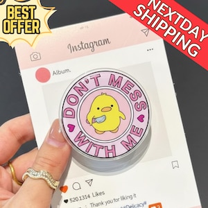 May include: A round, clear phone grip with a cartoon yellow chick holding a knife, surrounded by the text "DON'T MESS WITH ME" on a pink background. The grip is held over a mock Instagram post.