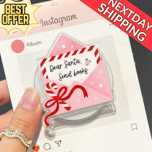 May include: A pink and red acrylic phone grip shaped like an envelope with the text "Dear Santa, Send books." The grip features a candy cane and bow design, with a clear base. The background includes Instagram-themed elements.