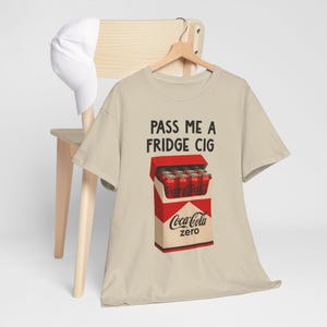 May include: Beige t-shirt with the text "PASS ME A FRIDGE CIG". The shirt features a graphic of a Coca-Cola Zero box filled with Coca-Cola cans. A white baseball cap hangs on a wooden chair.
