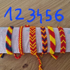 Può includere: Sei braccialetti dell'amicizia colorati con intricati motivi intrecciati in rosso, giallo e blu. Ogni braccialetto è esposto su un panno bianco, con i numeri da 1 a 6 scritti in blu sopra. I braccialetti hanno nappe.