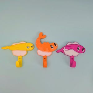 May include: Three wall hooks shaped like sushi pieces. The left hook is yellow, the center hook is orange, and the right hook is pink. Each sushi piece sits atop a white rice base and features a cartoon face with black eyes and a small hook.