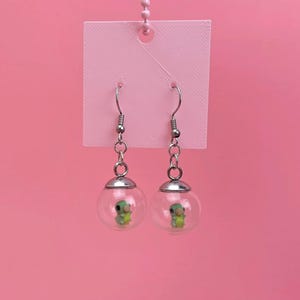 Peut inclure: Une paire de boucles d'oreilles pendantes avec des breloques sphériques en verre transparent, contenant chacune une petite figurine de chenille verte. Les breloques sont fixées à des crochets en métal argenté par de courtes chaînes argentées.