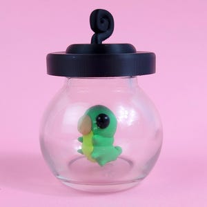 Peut inclure: Un petit bocal en verre transparent avec un couvercle et une poignée noirs, contenant un personnage de dessin animé vert et jaune. Le personnage a de grands yeux noirs et tient un objet jaune. Le bocal est posé sur un fond rose.