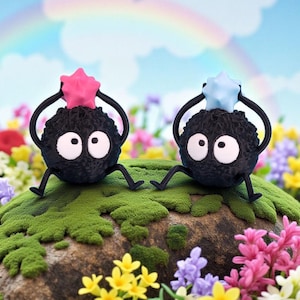 May include: Two black, round, animated figures with large white eyes and star-shaped headpieces, one pink and one blue, sit on a mossy rock. They have small black legs and arms. The background features a rainbow and colorful flowers.