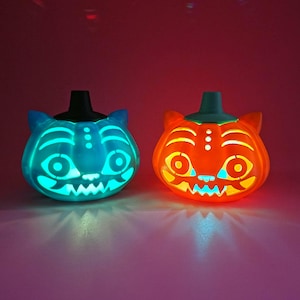 Pode incluir: Duas lanternas de abóbora em forma de gato com recortes brilhantes. A lanterna esquerda é azul com um caule preto, e a direita é laranja com um caule verde-azulado. Ambas apresentam traços faciais estilizados, incluindo olhos e dentes.