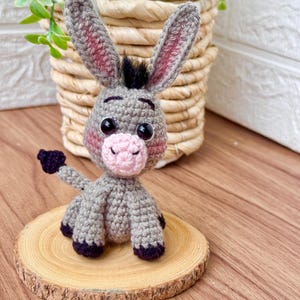 Może przedstawiać: Ręcznie robiona figurka osła na szydełku w kolorze szarym, różowym i czarnym. Osiołek ma duże, oklapnięte uszy z różowymi wnętrzami, różowy pysk i czarne oczy. Siedzi na małym drewnianym plastrze, prezentując szczegóły projektu amigurumi.