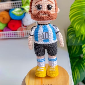 May include: A handcrafted crochet doll of a footballer with brown hair and a beard, wearing a light blue and white striped jersey with the number 10, black shorts, white socks with blue stripes, and yellow shoes, standing on a round wooden base.
