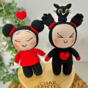 May include: Two handmade crochet dolls, one in red and one in black, with heart accents. The dolls have black hair and are on a wooden heart-shaped base. The base has the text "CHAR LOTTEE" on it. A red heart is attached to the top of the red doll.