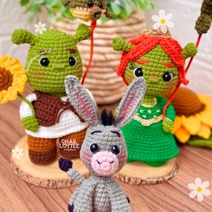 May include: Crocheted figures of Shrek, Fiona, Donkey, and a dragon. Shrek holds a sunflower, Fiona wears a crown, and the dragon is attached to a string. The figures are green, brown, grey, and red, with black eyes. The figures are on wooden slices.