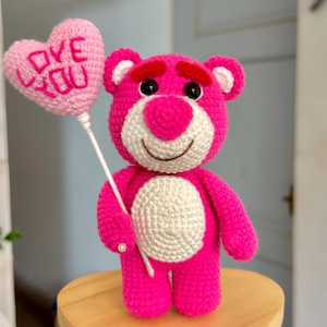 May include: A handmade pink crochet bear holding a pink heart-shaped balloon that says "LOVE YOU". The bear is standing on a wooden heart-shaped base with the text "CHAR LOTTEE #crochet".
