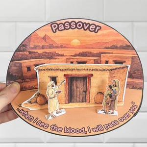 Pode incluir: Um artesanato em papel circular com o tema da Páscoa, com um fundo de paisagem desértica. A cena inclui uma casa, figuras e as palavras "Passover" e "...when I see the blood, I will pass over you." O artesanato é segurado por uma mão.