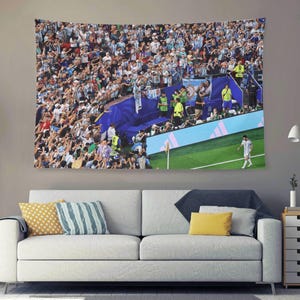 May include: A rectangular wall tapestry featuring a high-angle view of a crowded soccer stadium. A large crowd in light blue and white jerseys fills the stands, while a single player in a white jersey walks on the green grass near a blue barrier.
