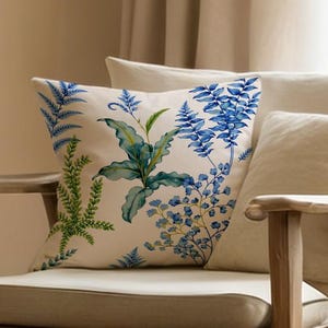May include: A wooden armchair with a beige cushion and backrest. A decorative pillow with a botanical print featuring blue and green leaves and flowers rests on the chair. Additional beige pillows provide support. The chair is positioned near a window with sheer curtains.