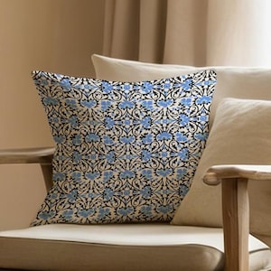 May include: A wooden armchair with a beige cushion and pillows. A decorative square pillow with a blue floral pattern rests on the chair. The chair is positioned in front of a beige curtain.