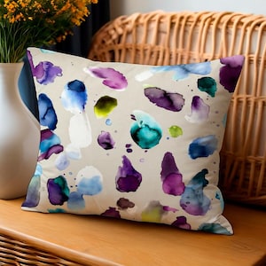 May include: A decorative pillow with a beige background and colorful watercolor splotches in shades of purple, blue, green, and white. The pillow is rectangular and appears to be made of a soft fabric. A white vase with yellow flowers is to the left.