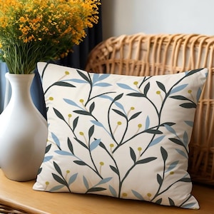 May include: Square decorative pillow with a cream background and a botanical pattern of dark green, light blue, and yellow leaves and berries. The pillow is placed on a wooden surface next to a white vase with yellow flowers.