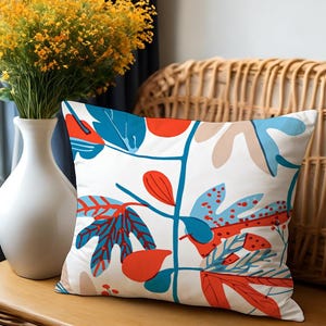 May include: Square decorative pillow with a white background and a colorful botanical design. The pattern features blue, red, and tan leaves and berries on a stem. The pillow is placed on a wooden surface.
