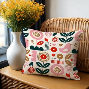 May include: Decorative throw pillow with a floral and bird pattern. The pillow features a cream background with pink, red, green, and brown geometric flowers, leaves, and a bird. The pillow is rectangular and sits on a wooden surface.