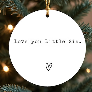 May include: A white, round Christmas ornament with the text "Love you Little Sis." in black, along with a small black heart. The ornament hangs from a gold string, set against a backdrop of a Christmas tree.
