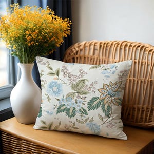 May include: A decorative square pillow with a floral pattern in shades of blue, green, and tan on a cream background. The pillow is placed on a wooden surface next to a white vase with yellow flowers and a wicker chair.