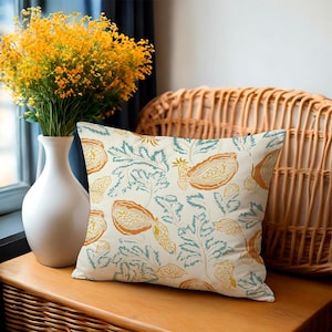 May include: A decorative square pillow with a cream background and a repeating pattern of orange and yellow fruit and teal leaves. The pillow is placed on a wooden surface next to a white vase with yellow flowers.