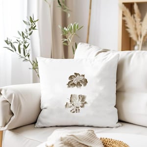 May include: White square throw pillow featuring two brown floral designs. The pillow is placed on a white sofa, with other pillows and decorative plants in the background. The floral pattern adds a touch of nature-inspired decor.