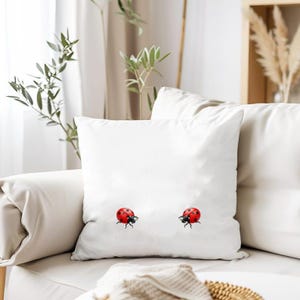 May include: White square throw pillow with two red ladybug designs. The ladybugs have black spots and legs. The pillow is on a white sofa with a neutral background.
