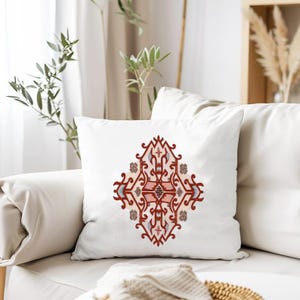 May include: White square throw pillow with a symmetrical, ornate design in shades of red, pink, and blue. The pattern is centered on the pillow, creating a decorative accent for home decor. The pillow is on a white sofa.