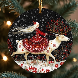 Peut inclure: Ornement de Noël rond représentant une femme en robe rouge chevauchant un renne blanc dans un paysage enneigé. L'ornement a un fond noir avec des flocons de neige et des accents floraux rouges. Une ficelle dorée est attachée pour l'accrocher.