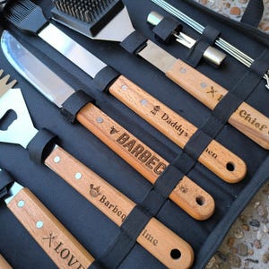 Puede incluir: Un estuche negro con cremallera contiene un juego de herramientas para barbacoa. El juego incluye una espátula, un cuchillo, un cepillo y brochetas con mangos de madera. Algunos mangos están grabados con texto como "Daddy's BBQ" y "Chief".