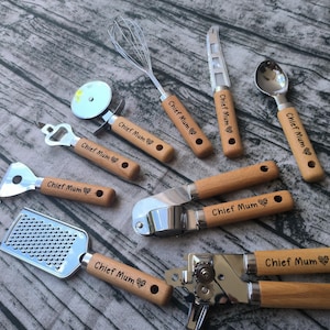 Puede incluir: Un conjunto de utensilios de cocina con mangos de madera y componentes de acero inoxidable. Los utensilios incluyen un cortador de pizza, un abridor de botellas, un rallador, un batidor, un cuchillo, una cuchara, un prensador de ajos y un abrelatas. Cada mango está grabado con "Chief Mum".
