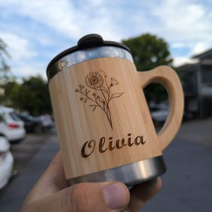 May include: A personalised travel mug made of bamboo and stainless steel. The mug features a floral design and the name "Olivia" engraved on the bamboo. It has a black lid and a handle. The mug is designed for hot or cold beverages.