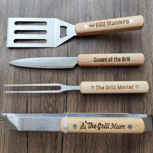Puede incluir: Un juego de cuatro utensilios para barbacoa de acero inoxidable y madera. El juego incluye una espátula, un cuchillo, un tenedor y unas pinzas. Los mangos de madera están grabados con las frases "BBQ Masters", "Queen of the Grill", "The Grill Master" y "The Grill Mum".