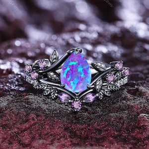 Könnte beinhalten: Ein schwarzer Ring mit einem birnenförmigen, irisierenden blau-violetten Edelstein. Der Ring hat ein florales Design mit kleinen rosa Edelsteinen und Blattakzenten. Der Ring liegt auf einer dunklen, strukturierten Oberfläche.