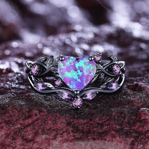 Puede incluir: Un anillo negro con una piedra central de ópalo iridiscente en forma de corazón. El anillo presenta un diseño floral con pequeñas piedras preciosas rosas y detalles de hojas. El ópalo muestra tonos de azul y morado.