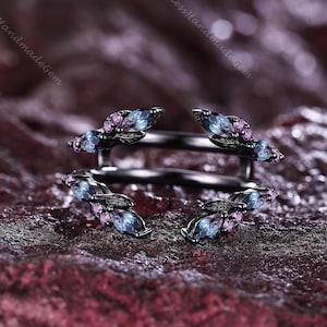 May include: A black metal ring featuring a double-band design with an open front. The bands are adorned with marquise-cut blue stones and small round purple stones arranged in a leaf-like pattern along the sides of the ring.
