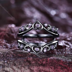 Puede incluir: Un protector de anillo negro con un diseño en forma de corazón y pequeñas piedras preciosas moradas. El protector de anillo está diseñado para usarse con otro anillo. El protector de anillo tiene un estilo vintage.