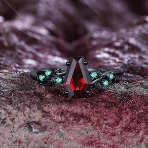 May include: A black ring with a large, rectangular, deep red gemstone as the centerpiece. The ring features small green gemstones and a decorative vine design. The words "TimelessHandmadeGem" are visible in the background.