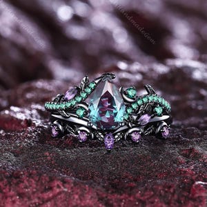 May include: A black ring set with a large, pear-shaped, teal-colored gemstone. The ring is accented with smaller green and purple gemstones. The band has a floral design. The words "TimelessHandmadeGem" are in the background.