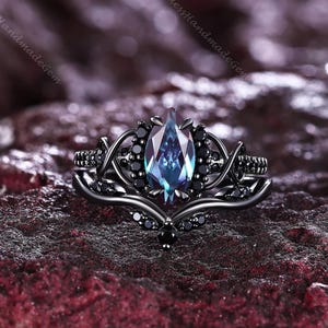 May include: A black metal ring featuring a central marquise-cut stone with blue and purple color-shifting hues. The band is designed with intricate vine-like metalwork and is accented with small, round, black stones set along the branches.