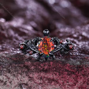 Puede incluir: Un anillo de metal negro con una piedra central ovalada de ópalo multicolor. El anillo presenta un diseño de tortuga con detalles intrincados y pequeñas gemas rojas en los lados. El anillo está sobre un fondo oscuro y texturizado.