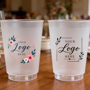 May include: Two frosted plastic cups featuring custom designs. The left cup has a pink circular floral wreath with the text YOUR LOGO HERE. The right cup displays a black rectangular frame with leaf accents and the text YOUR LOGO HERE.