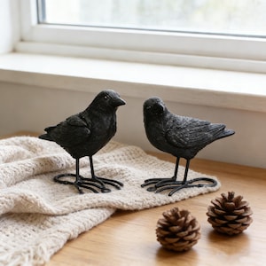 May include: Two black decorative crow figurines with detailed feather textures and long, thin legs. They are perched on a beige knitted blanket, with two pine cones nearby. The crows are placed on a wooden surface near a window.