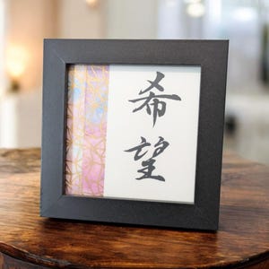 以下が含まれることがあります： 白い背景に日本の書道が施された正方形の黒いフォトフレーム。 書道は「希望」と読みます。 フレームには、金色のアクセントが付いたカラフルな抽象的なデザインも施されています。