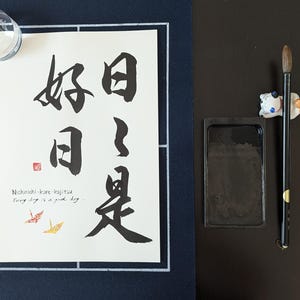 以下が含まれることがあります： 日本語の文字と「Every day is a good day」という英語のフレーズが書かれた額入りの書道作品。筆、インク皿、セラミック製の筆置きが含まれています。 作品は紺色のマットで額装されています。
