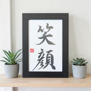 May include: Black framed art print with Japanese calligraphy. The artwork features two characters in black ink on a white background, with a red stamp in the lower left corner. Two small potted succulents flank the framed print.